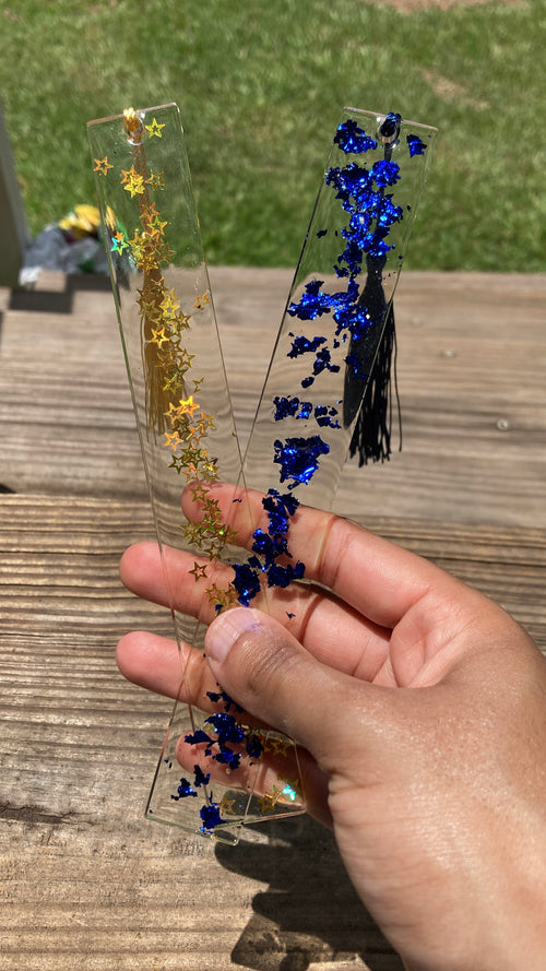 A hand holding two clear rectangular resin bookmarks or tags outdoors on a wooden table, one with gold glitter stars and the other with blue glitter flowers and black tassel.